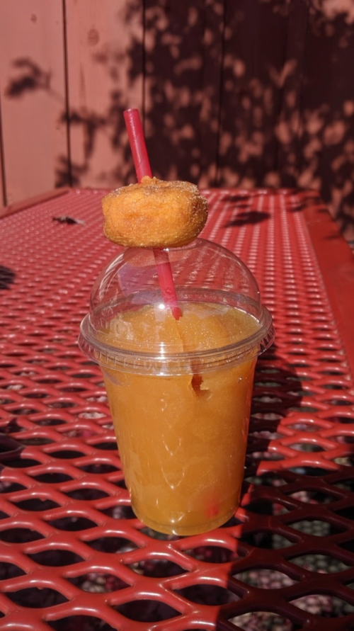 A clear cup of orange slush topped with a small donut, resting on a red metal table.