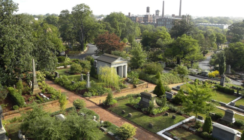 Aerial view of a serene cemetery with lush greenery, pathways, and monuments, set against an industrial backdrop.