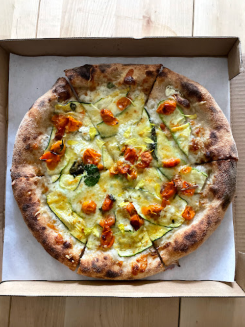A freshly baked pizza topped with yellow squash, cherry tomatoes, and melted cheese in a pizza box.