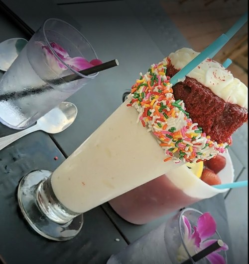 A tall milkshake topped with whipped cream and sprinkles, alongside a glass of water and a colorful dessert.