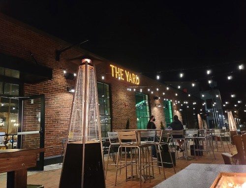 Outdoor seating area at night with string lights and a heater, featuring a brick building with "THE YARD" sign.