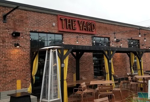 Exterior of a restaurant called "The Yard," featuring a brick facade, outdoor seating, and string lights.