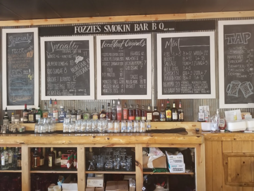 Chalkboard menu at Fozzie's Smokin Bar B Q, featuring specials, food items, and drink options. Rustic wooden bar below.