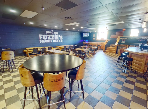Interior of Fozzie's Smokin Bar-B-Q, featuring tables, chairs, and a cozy atmosphere with a blue wall and TV.