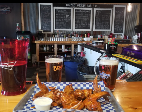 A table with chicken wings and two beers in front of a bar with a chalkboard menu in the background.
