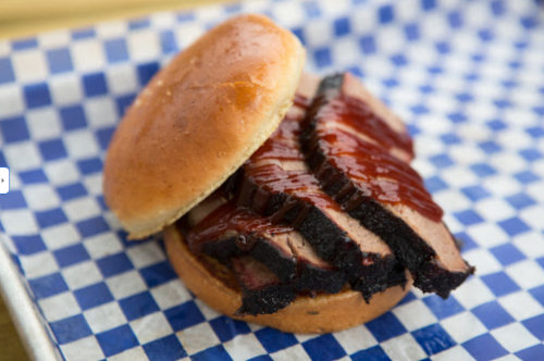 A close-up of a barbecue brisket sandwich with sauce on a bun, served on a blue and white checkered paper.