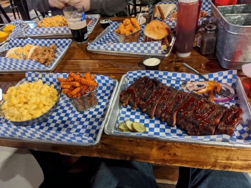 A table with trays of barbecue ribs, macaroni and cheese, sweet potato fries, and a burger, all on checkered paper.