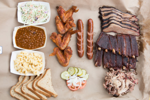 A spread of barbecue featuring wings, ribs, sausages, baked beans, coleslaw, macaroni, and sliced bread.