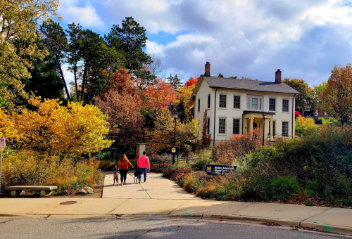 Two people walk towards a historic house surrounded by colorful autumn trees and a cloudy sky.