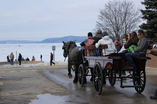 A horse-drawn carriage carries passengers along a snowy lakeside, with people walking on the shore in the background.