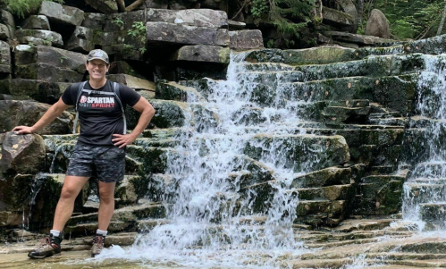 A person stands beside a cascading waterfall, surrounded by rocky terrain and lush greenery.