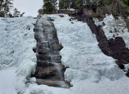 A frozen waterfall cascading down a rocky cliff, surrounded by snow and trees.