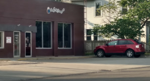 A red car parked beside a modern building with large windows and a sign, surrounded by greenery.