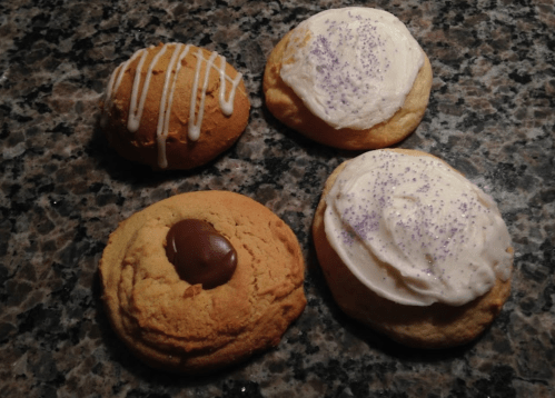 Four cookies on a granite surface: two with frosting and sprinkles, one with chocolate, and one drizzled with icing.