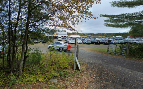 A gravel entrance with a white sign leads to a large parking area filled with cars, surrounded by trees.