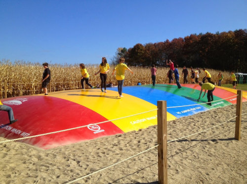 Children and adults play on a colorful inflatable bounce pad in a cornfield under a clear blue sky.