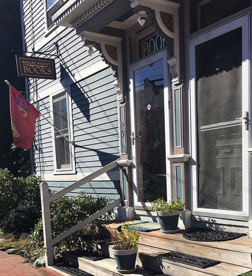 A quaint bookstore entrance with a sign reading "Sheaf Street Books," surrounded by potted plants and a welcoming porch.