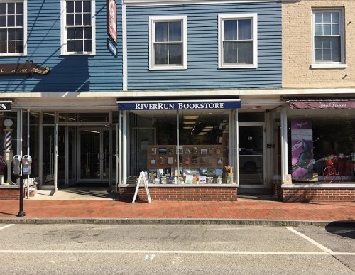 A quaint bookstore with a blue facade, featuring a display of books in the window and a sign reading "Riverrun Bookstore."