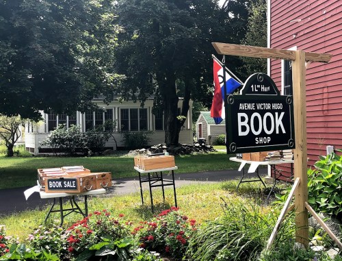 A quaint bookshop with a sign reading "Book Sale" and tables displaying books, surrounded by greenery and a house in the background.