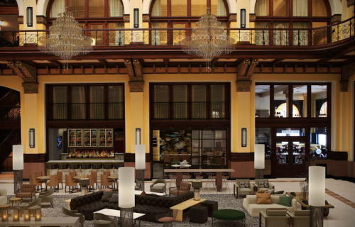 Elegant hotel lobby featuring chandeliers, modern seating, and a warm color palette with a bar area in the background.