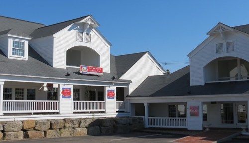 Two white buildings with gray roofs, featuring signs and a stone wall in front, under a clear blue sky.