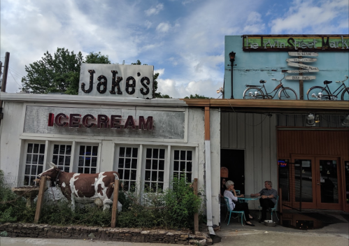 A quaint ice cream shop named "Jake's" with a cow statue outside, next to a building with bicycles on the roof.