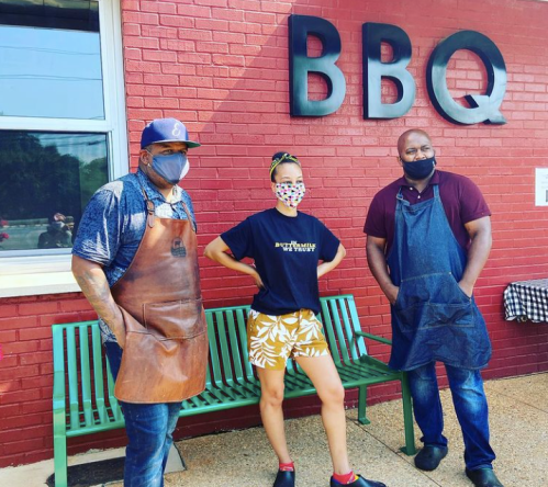 Three people in aprons stand outside a BBQ restaurant, wearing masks and posing in front of a red wall with "BBQ" signage.
