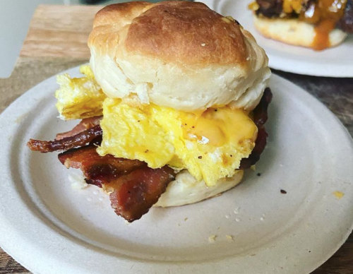 A biscuit sandwich with crispy bacon, scrambled eggs, and melted cheese on a paper plate.