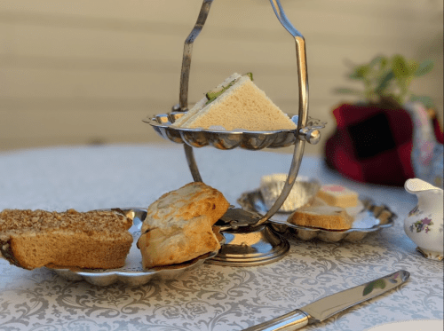 A tiered serving tray with sandwiches, scones, and bread on a table set with a floral tablecloth and a small plant.