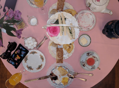 A beautifully arranged tea table with sandwiches, pastries, a rose, and drinks on a pink tablecloth.