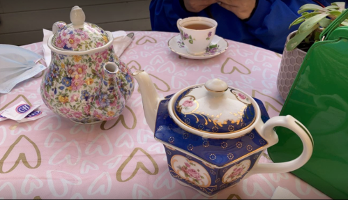A floral teapot and a blue patterned teapot on a heart-patterned tablecloth, with a cup of tea in the background.