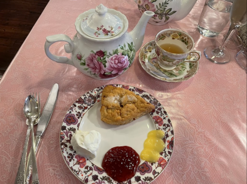 A floral teapot and cup beside a plate with a scone, whipped cream, jam, and lemon curd on a pink tablecloth.
