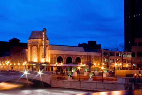 A historic building illuminated at dusk, reflecting on a river with city lights in the background.