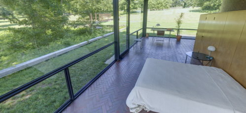 A modern bedroom with large glass windows overlooking a green landscape, featuring a bed and minimalist furniture.