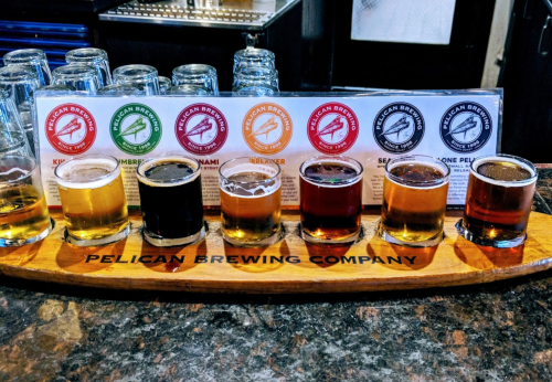 A wooden flight board with seven small beer glasses in various colors, alongside a menu from Pelican Brewing Company.