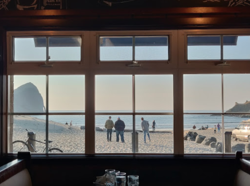 View through a restaurant window showing people on a beach with ocean waves and rocky cliffs in the background.