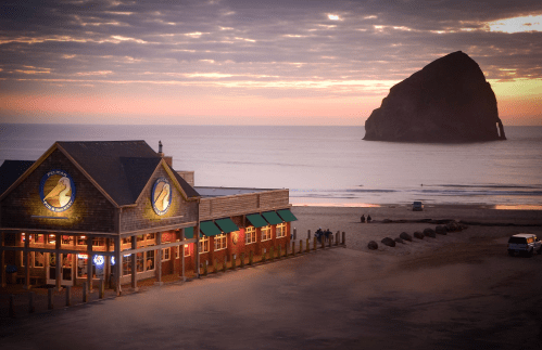 A beachside restaurant at sunset, with a large rock formation in the ocean and people walking along the shore.