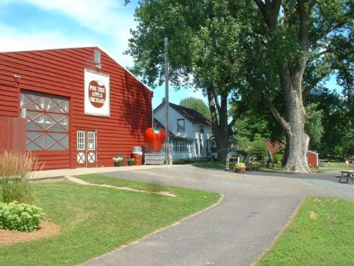 A red barn and white farmhouse are seen along a path, surrounded by green grass and trees. A large apple sculpture is nearby.