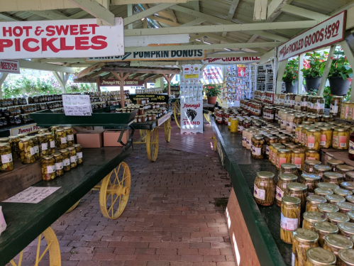 A market stall filled with jars of pickles and other goods, with signs for hot and sweet pickles and pumpkin donuts.