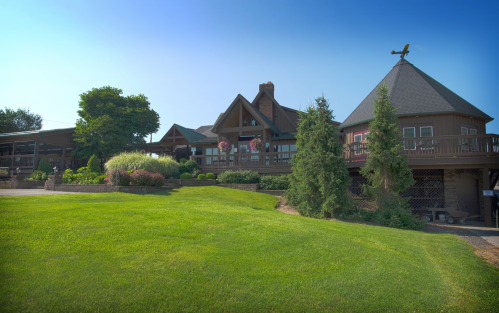 A large wooden lodge surrounded by lush green grass and trees under a clear blue sky.