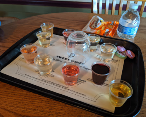 A tray with various small cups of sweet wine, a glass, and a water bottle, set on a table.