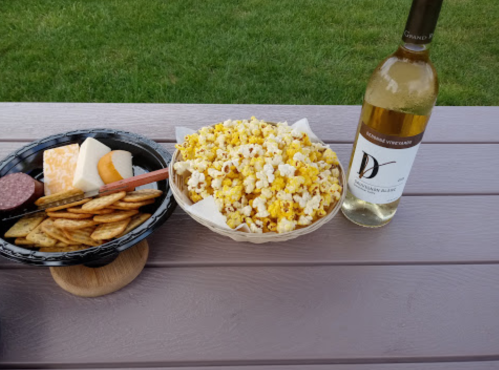 A bowl of popcorn, a plate of snacks, and a bottle of white wine on a wooden table outdoors.