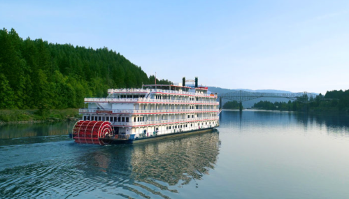 A large riverboat with a paddle wheel sails on a calm river, surrounded by lush green trees and distant hills.