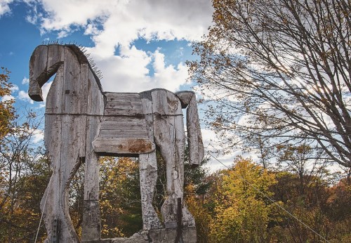 A large wooden horse sculpture stands against a backdrop of trees and a cloudy sky.
