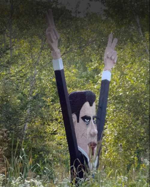 A large, stylized statue of a man with dark hair and raised hands, surrounded by trees in a natural setting.