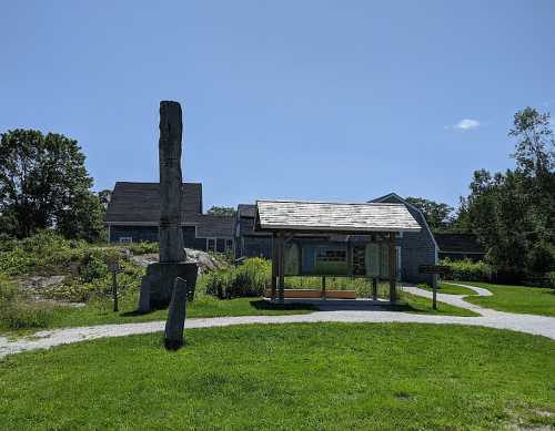 A grassy area featuring a totem pole, a shelter, and buildings under a clear blue sky.