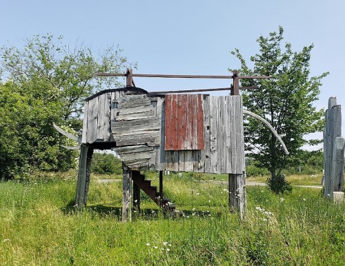 A wooden structure resembling an elephant, with stairs and a rustic design, set in a grassy area with trees.