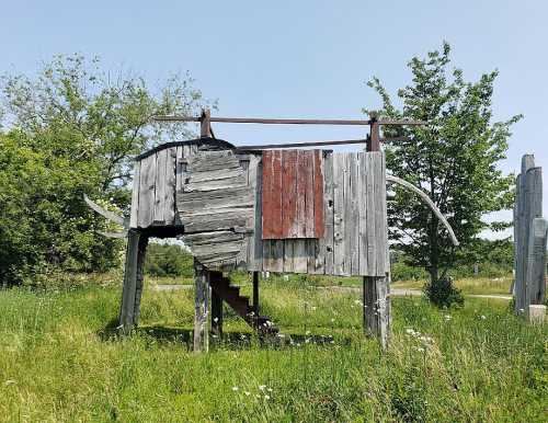 A wooden structure resembling an elephant, with stairs and a rustic design, set in a grassy area with trees.