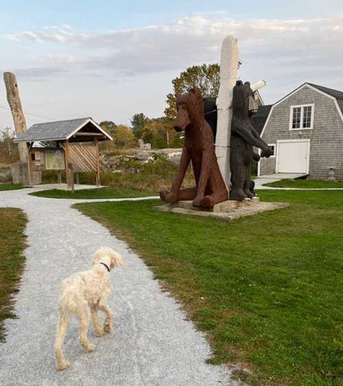 A dog walks along a path near wooden bear sculptures and a rustic building in a grassy area.