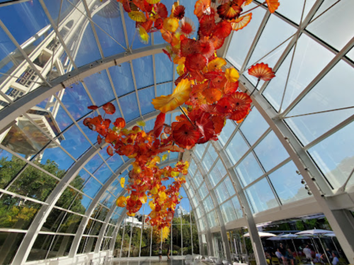 Colorful glass flowers in shades of orange and red hang from a glass ceiling, with a blue sky visible above.